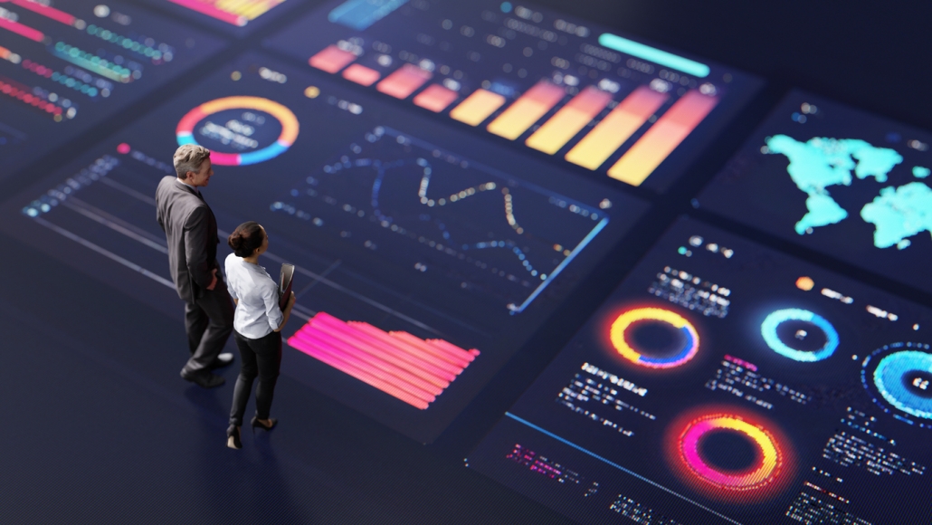 Two businesspeople stand on a dark floor displaying large, colorful digital graphs and charts—bar graphs, line charts, circular diagrams, and a world map—illustrating data analysis as one of the key Pillars of Successful Branding.