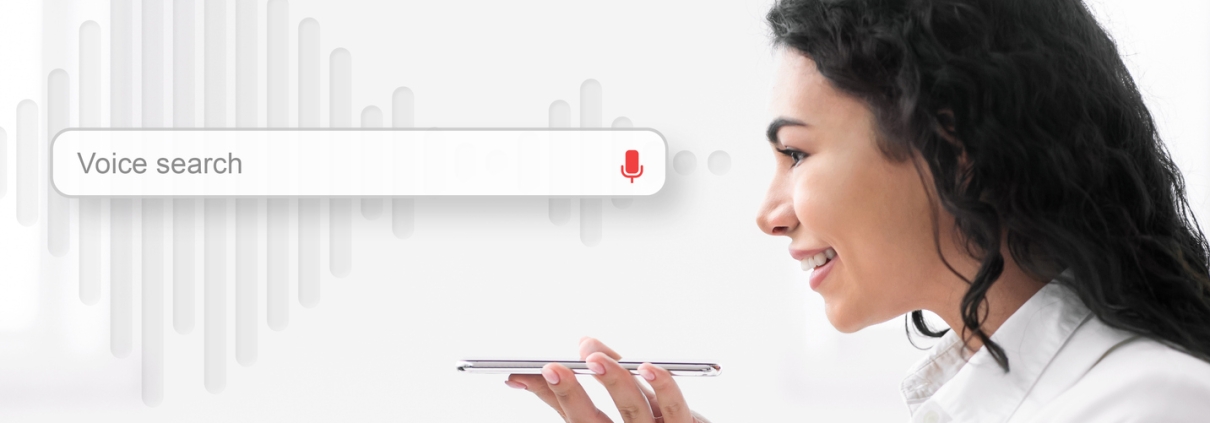 A woman smiles while holding a smartphone and using voice search. A graphic of a sound wave and a search bar with a microphone icon and the words Voice Search SEO are displayed next to her.