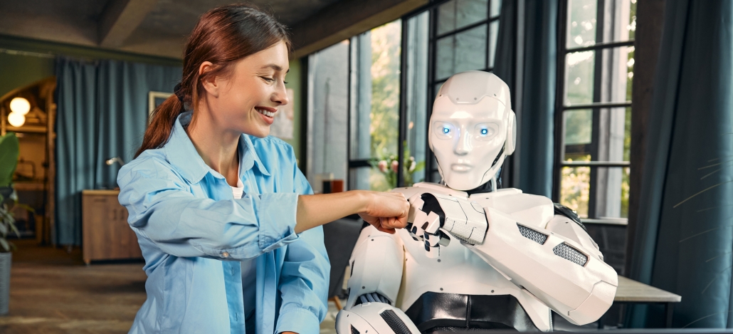 A smiling woman in a blue shirt fist bumps a white humanoid robot, showcasing the friendly side of Artificial Intelligence In Digital Marketing, while sitting indoors in a modern, sunlit room with large windows and blue curtains.