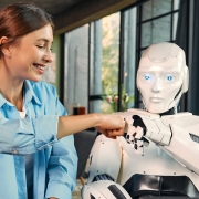 A smiling woman in a blue shirt fist bumps a white humanoid robot, showcasing the friendly side of Artificial Intelligence In Digital Marketing, while sitting indoors in a modern, sunlit room with large windows and blue curtains.