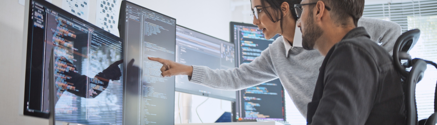 Two software developers look at computer monitors displaying code. One points at the screen while the other uses a mouse, both appearing focused and engaged in discussion about implementing an AI powered search feature in a bright office space.