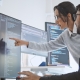 Two software developers look at computer monitors displaying code. One points at the screen while the other uses a mouse, both appearing focused and engaged in discussion about implementing an AI powered search feature in a bright office space.