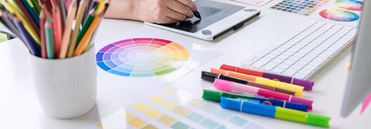 A person uses a digital drawing tablet at a desk covered with color swatches, a printed color wheel, colored pencils, highlighters, and a computer keyboard, reflecting a creative workspace focused on graphic design and branding strategy.