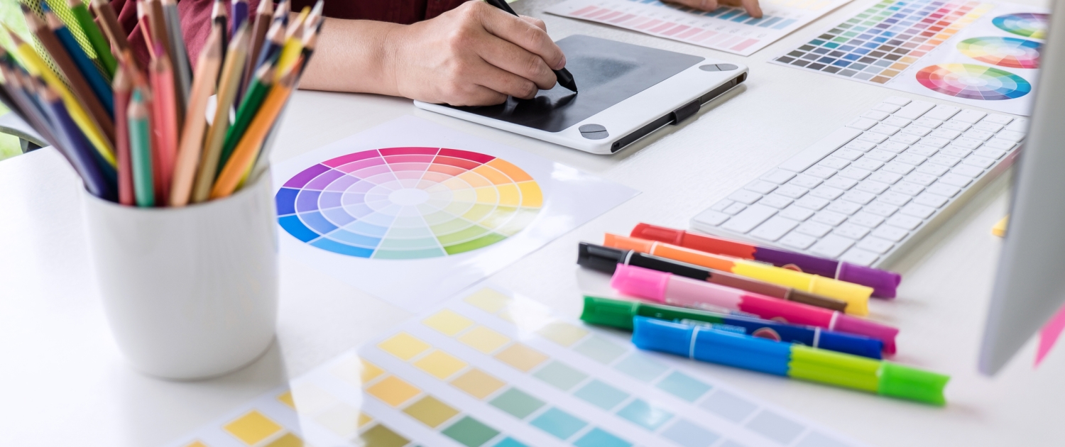 A person uses a digital drawing tablet at a desk covered with color swatches, a printed color wheel, colored pencils, highlighters, and a computer keyboard, reflecting a creative workspace focused on graphic design and branding strategy.