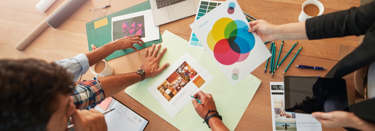 Three people gather around a wooden table with papers, a color chart, a laptop, pens, and a tablet, collaborating on a creative design project and discussing branding strategy. One person holds a sheet with a color wheel.