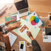 Three people gather around a wooden table with papers, a color chart, a laptop, pens, and a tablet, collaborating on a creative design project and discussing branding strategy. One person holds a sheet with a color wheel.