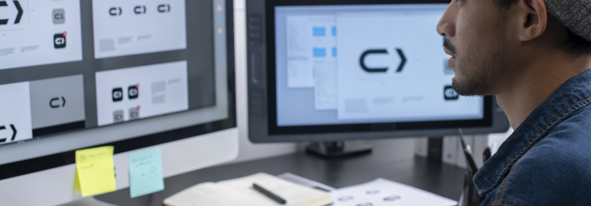 A man wearing a beanie works at a desk with two monitors, reviewing logo designs. Papers, sticky notes, and a notebook are on the desk. The screens display variations of a black and white arrow logo.