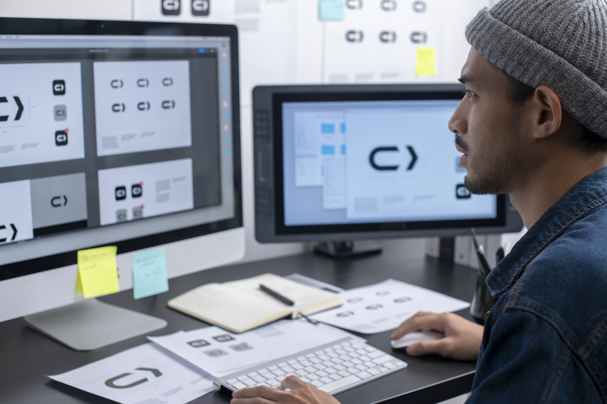 A man wearing a beanie works at a desk with two monitors, reviewing logo designs. Papers, sticky notes, and a notebook are on the desk. The screens display variations of a black and white arrow logo.
