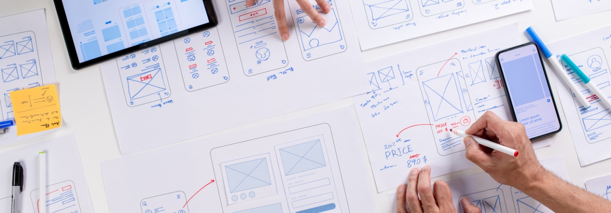 People collaborating on a table covered with wireframes, sketches, and digital devices, planning mobile app designs to address challenges like a website not generating leads, using paper prototypes, a tablet, a smartphone, and assorted pens.