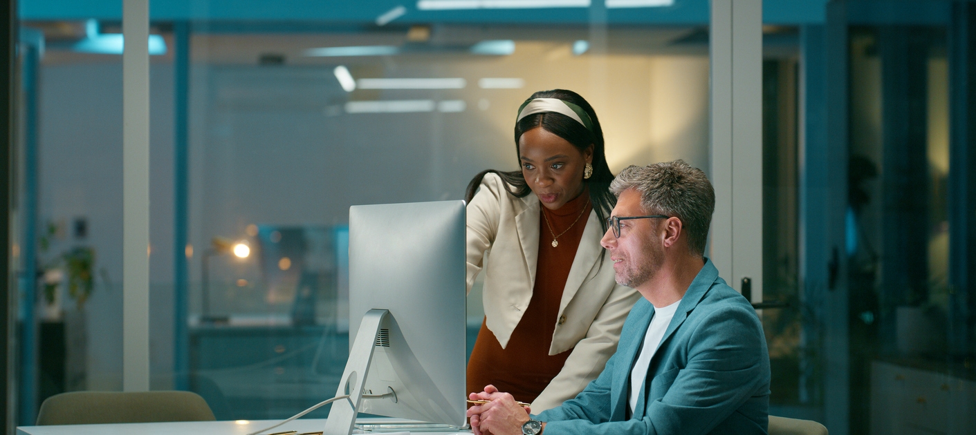Two colleagues in a modern office work late at a computer. A woman stands and gestures at the monitor, discussing why their website is not generating leads, while a man sits attentively. Papers and a notebook are on the desk.