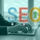 A person in business attire types on a laptop, with large translucent letters SEO and an upward arrow overlaying the image, symbolizing search engine optimization and growth.
