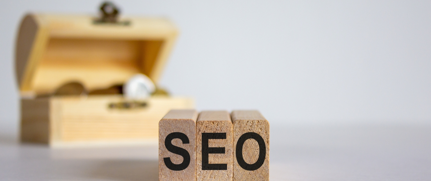 Three wooden blocks with the letters SEO are arranged in the foreground, while an open wooden treasure chest with coins inside is blurred in the background, symbolizing the value of SEO.