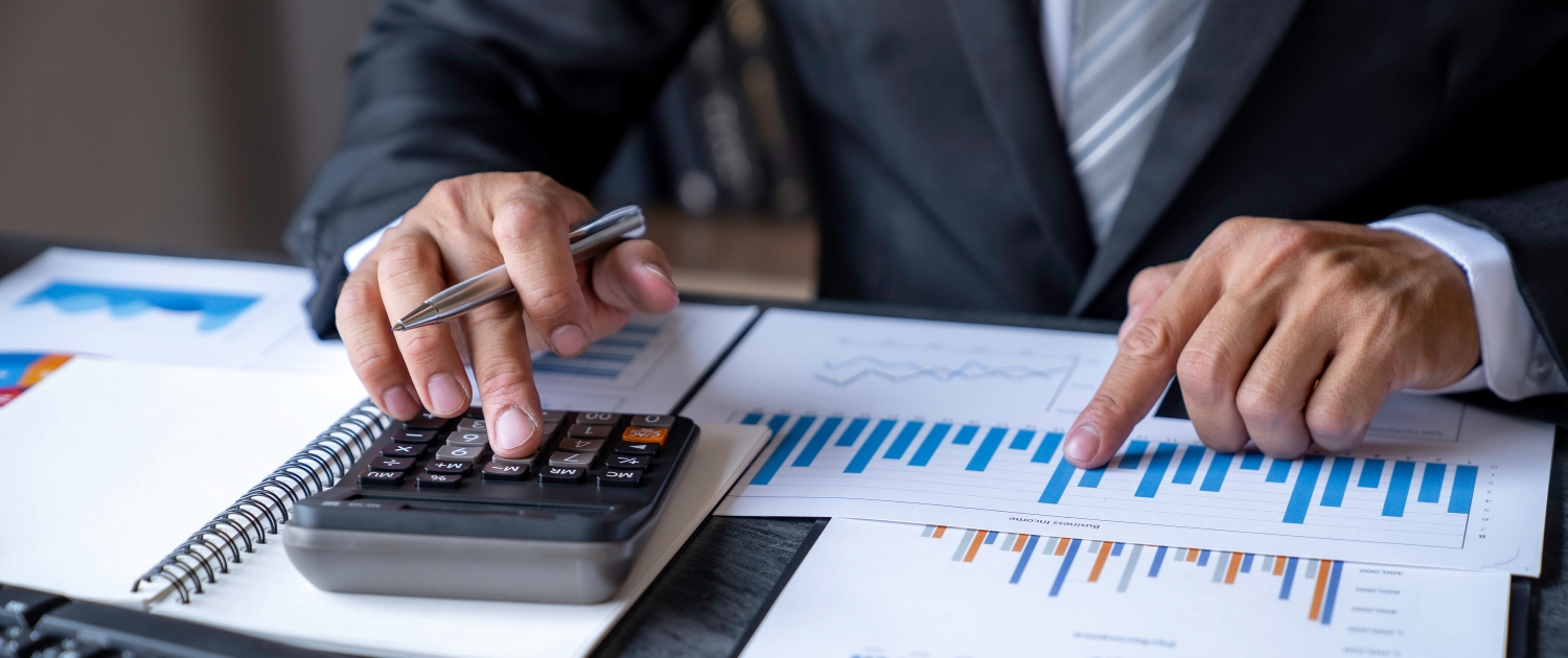 A person in a suit uses a calculator and points to a bar graph on a paper, surrounded by documents and charts on a desk, suggesting financial analysis or business planning.