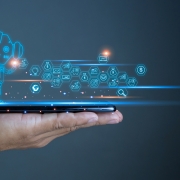 A hand holds a smartphone with blue holographic icons and a robot figure projected above it, symbolizing artificial intelligence and technology.