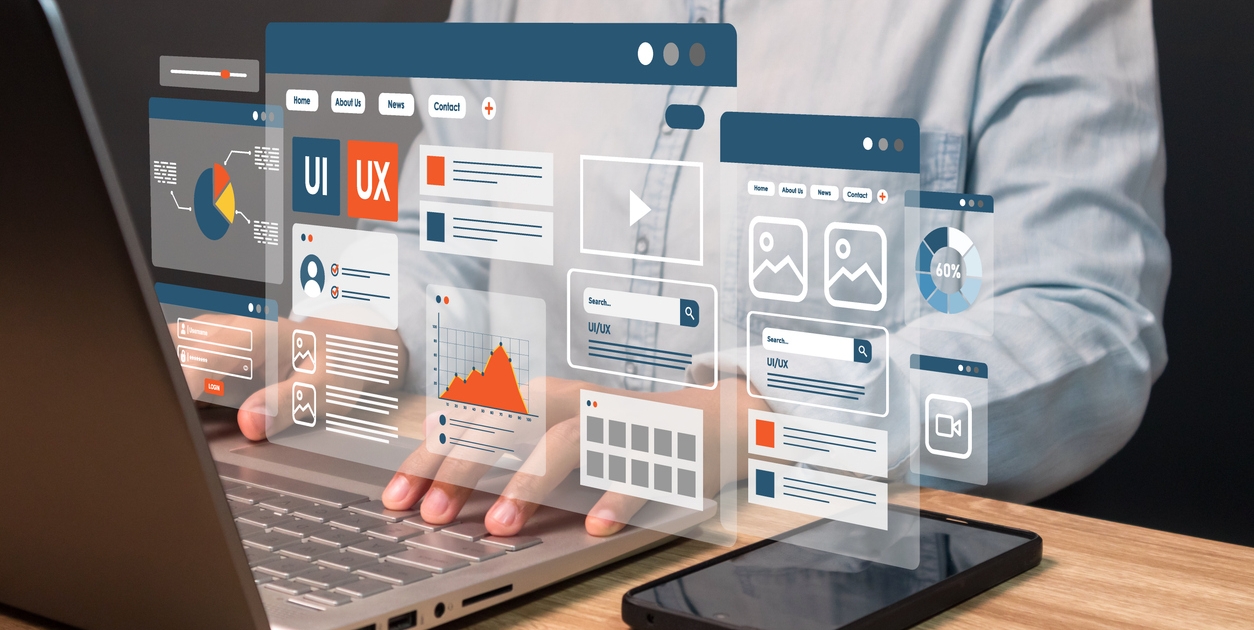 A person uses a laptop with floating digital UI and UX interface elements, such as charts, graphs, and web pages, while a smartphone lies on the wooden desk beside them.