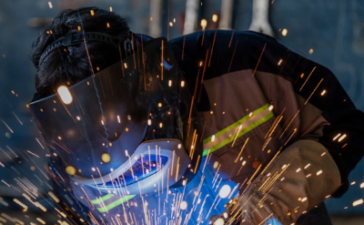 A person wearing a welding helmet works on a project, surrounded by bright flying sparks.